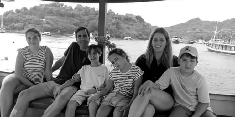 A family of six, including Fernando Martín Carreras, his wife, and their four children, posing happily on the wooden deck of the traditional pinisi schooner KM Putri Sakinah. They are smiling against the backdrop of the arid, rolling hills of Labuan Bajo and a clear blue sky on December 26, 2025.
