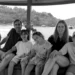 A family of six, including Fernando Martín Carreras, his wife, and their four children, posing happily on the wooden deck of the traditional pinisi schooner KM Putri Sakinah. They are smiling against the backdrop of the arid, rolling hills of Labuan Bajo and a clear blue sky on December 26, 2025.