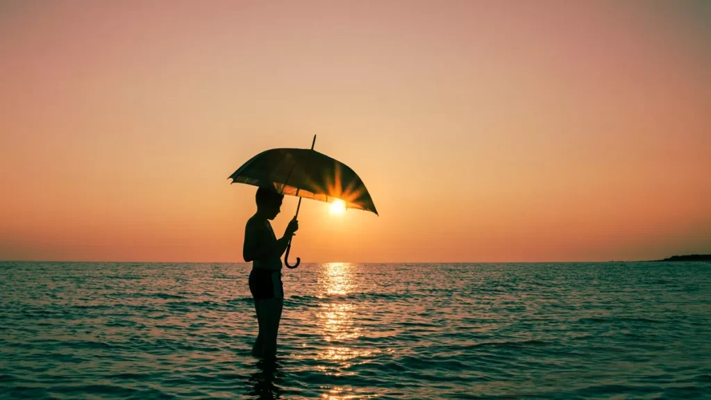 Bali Beach Safety During Rainy Season: 7 Realistic Tips for Travelers & Expats 2 Illustration photo of a foreign tourist enjoying the sunset on a Bali beach while carrying an umbrella during the rainy season (Hey Bali)