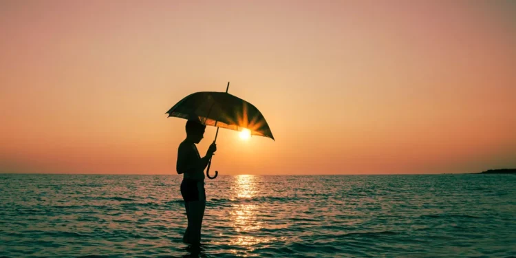 Bali Beach Safety During Rainy Season: 7 Realistic Tips for Travelers & Expats 37 Illustration photo of a foreign tourist enjoying the sunset on a Bali beach while carrying an umbrella during the rainy season (Hey Bali)