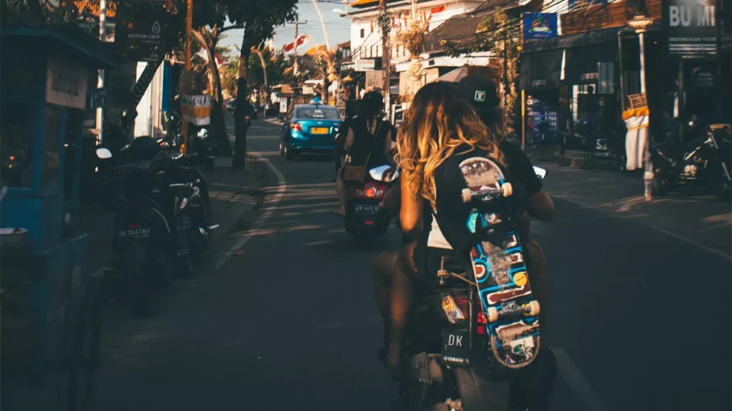 Bali's Most Famous Destinations Aren't Losing Their Magic. They're Becoming Victims of Their Own Success. 4 Photo of the atmosphere on a street in Bali shows a Caucasian woman riding a motorbike while carrying a skateboard.