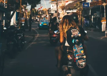 Photo of the atmosphere on a street in Bali shows a Caucasian woman riding a motorbike while carrying a skateboard.
