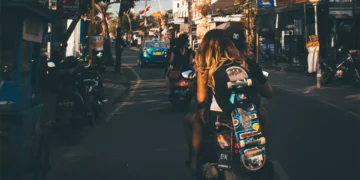 Photo of the atmosphere on a street in Bali shows a Caucasian woman riding a motorbike while carrying a skateboard.