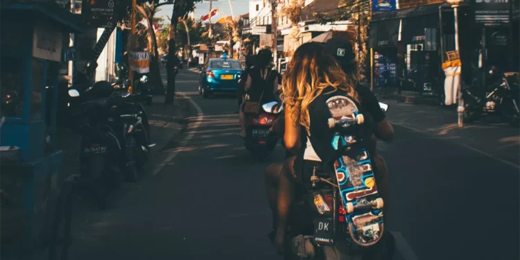 Photo of the atmosphere on a street in Bali shows a Caucasian woman riding a motorbike while carrying a skateboard.