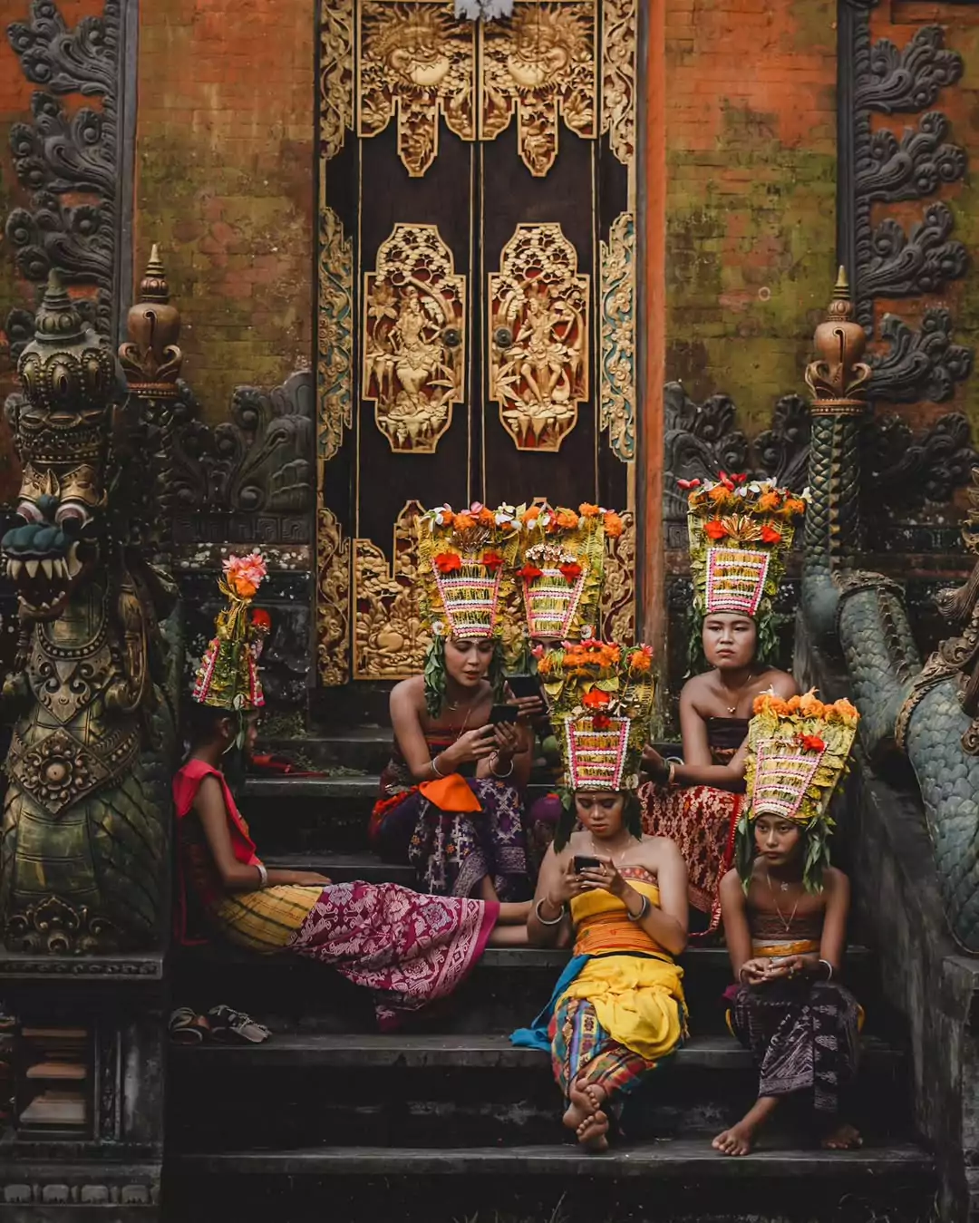 Six Balinese teenage girls in traditional attire sit together at Puri Ubud, engrossed in their smartphones—a contrast between cultural heritage and modern digital influence.