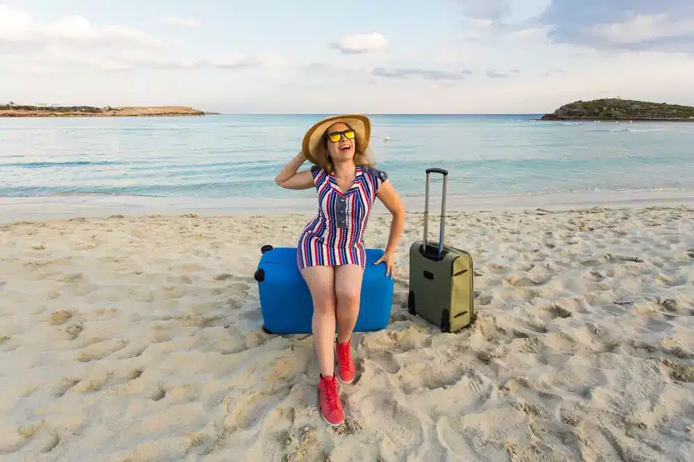 a female tourist posing on the beach with 2 suitcases behind her and a beautiful sea background