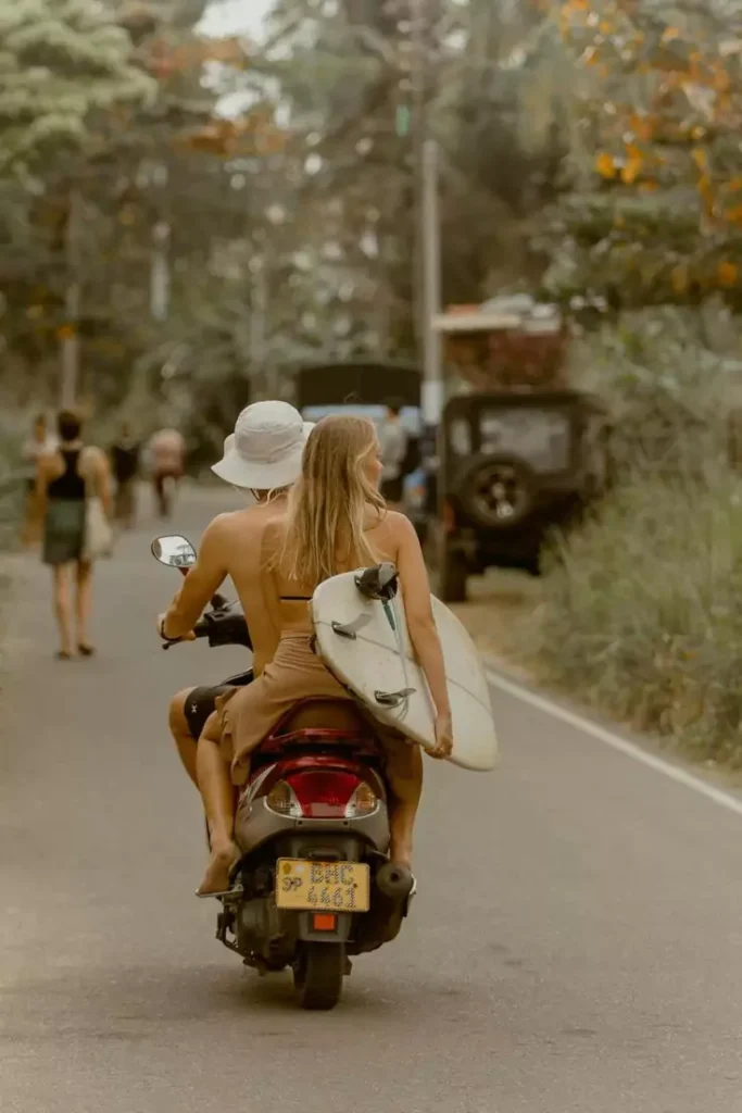 Photo of a couple of Caucasians riding a motorcycle while carrying a surfboard without wearing a helmet and without proper clothing - Bali tourist problems