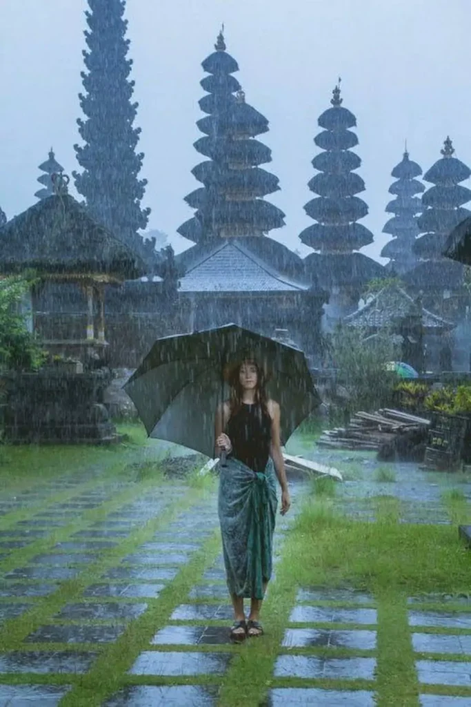A photo of a woman at a temple in Bali in the middle of heavy rain. The woman appears to be standing with an umbrella in the heavy rain.