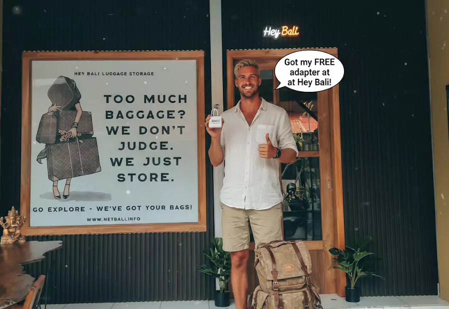 A blonde male traveler from Australia smiles contentedly in front of 'Hey Bali Luggage Storage', holding his newly acquired Free Bali adapter. His travel backpack lies to the side, showing how convenient it is to prepare for a trip to Bali.