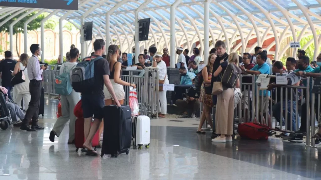 Bali Airport Scramble for transportation, illustrating the high pressure from drivers to new arrivals. The image shows a crowded arrival area at I Gusti Ngurah Rai International Airport (DPS), with tourists pulling luggage past groups of waiting local people (likely drivers or touts). This visual emphasizes why knowing the phrase "Saya sudah pesan online, thank you" is essential for travelers to avoid being swarmed and to secure a reliable Bali airport transfer to Ubud or other destinations.