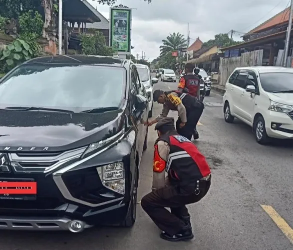 Ubud Parking Fine enforcement action being taken against an illegally parked black car (Mitsubishi Xpander). Two local police/traffic officers, wearing uniforms marked 'POLIS', are inspecting the vehicle, highlighting the strict new regulations against illegal parking scams and roadside parking. This relates to potential driver parking fee scams often mentioned by non-official drivers to tourists.