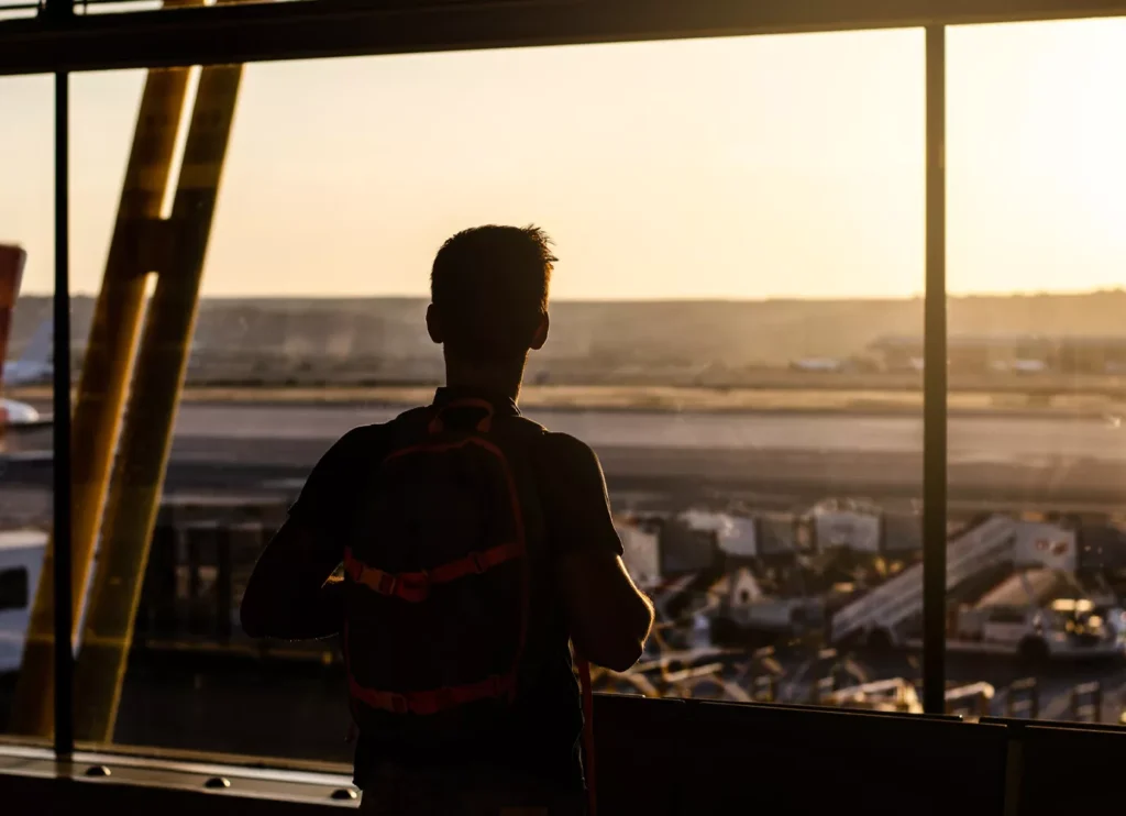 A silhouette of a male traveler with a backpack looking out of a large airport terminal window at sunset. The golden light illuminates the tarmac, airport service vehicles, and the runway beyond. This image captures the moment of arrival and anticipation before a traveler organizes their Bali airport transfer to their final destination.
