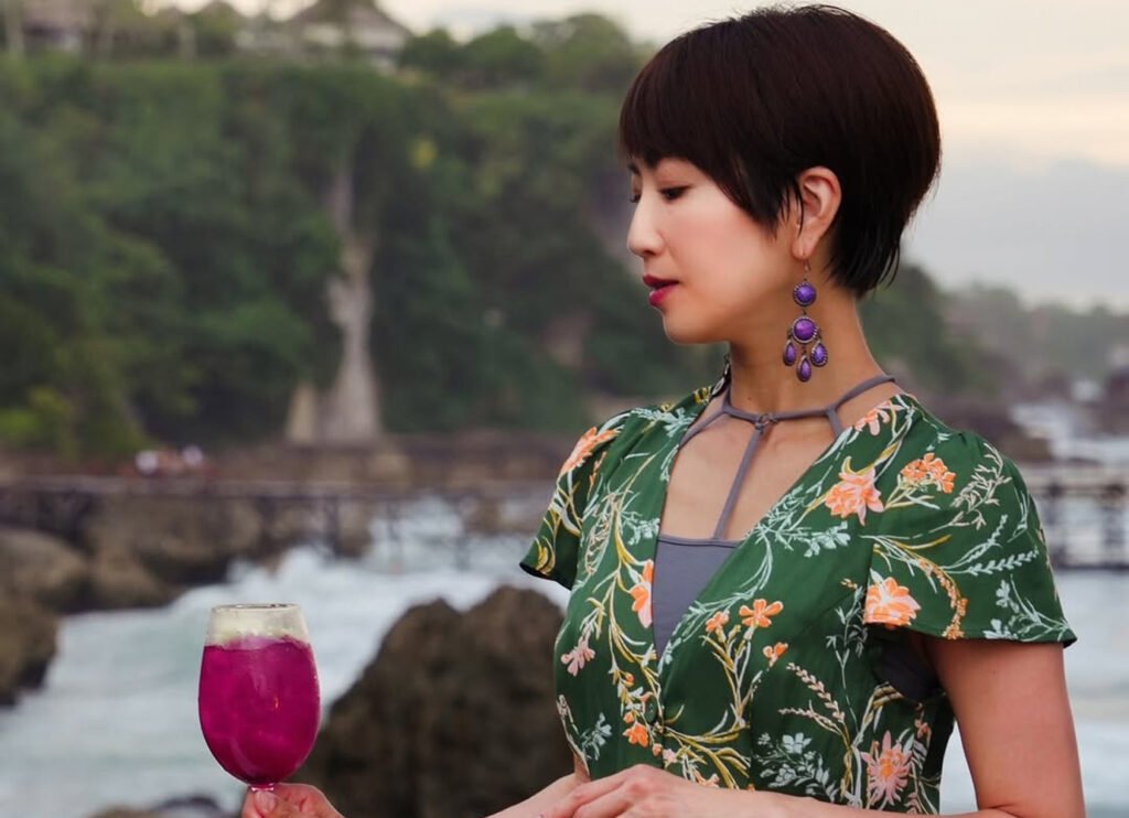 A close-up portrait of a woman with short dark hair, wearing a green floral dress and large purple dangle earrings. She is holding a glass of bright purple cocktail (likely dragon fruit) against a blurred background of green foliage and a rocky coastal view with wooden walkways. The image emphasizes a relaxed, stylish holiday moment.