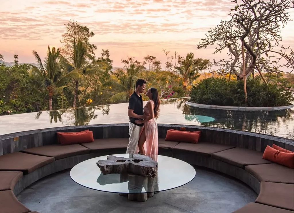 Bali Airport to Jimbaran Transfer successful arrival at the Rimba Jimbaran by Ayana resort. A romantic photo of a couple embracing at sunset next to an infinity pool and a circular lounge area.