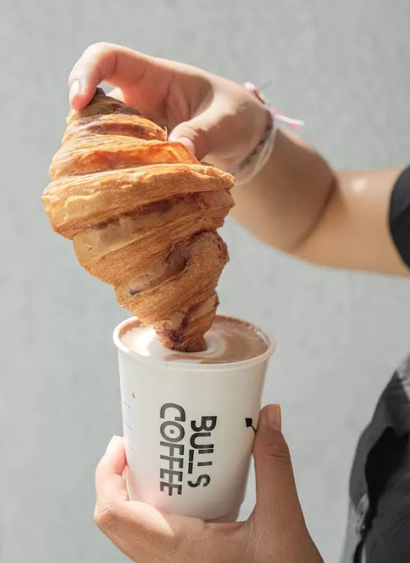 Hand dipping a crispy croissant into a cup of hot chocolate or coffee from Bull's Coffee