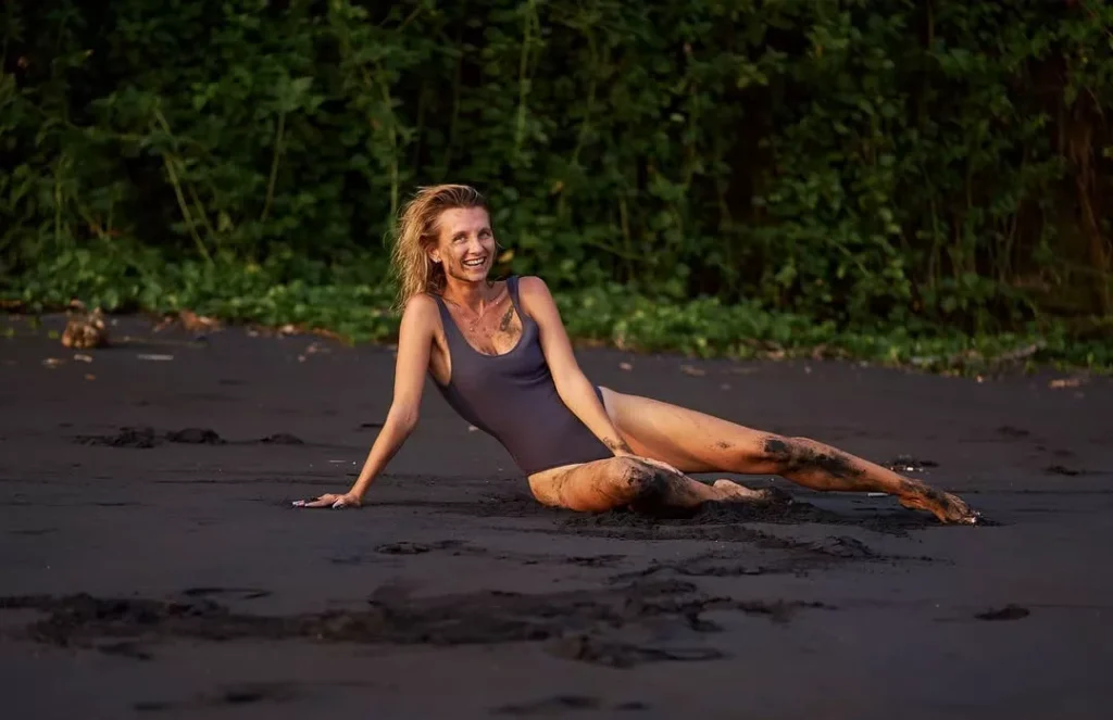 Happy woman in a dark swimsuit relaxing on Bali's black volcanic sand, covered in dirt and smiling brightly, suggesting an authentic, off-the-checklist experience away from the polished resort look. Alt: The real answer to is Bali overrated lies in abandoning the algorithmic loop for true discovery.