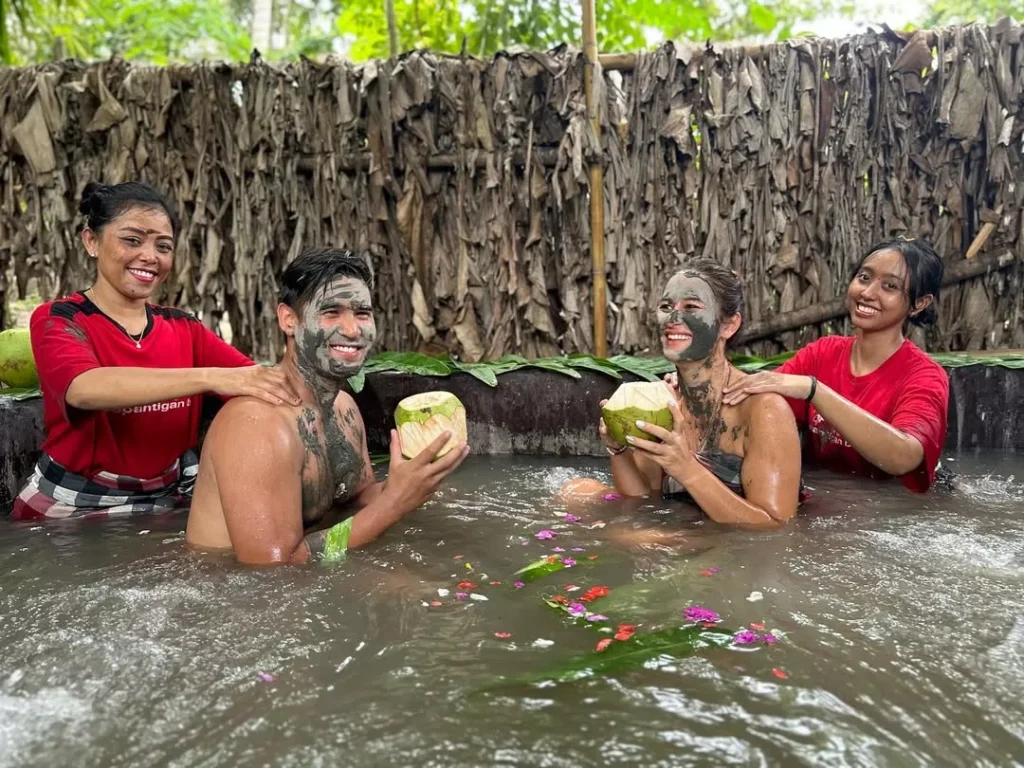 Smiling couple enjoying a unique mud bath and massage with local hosts in Bali, holding fresh coconuts. This authentic and joyous interaction provides the ultimate answer: Bali is not boring when you truly connect with it.