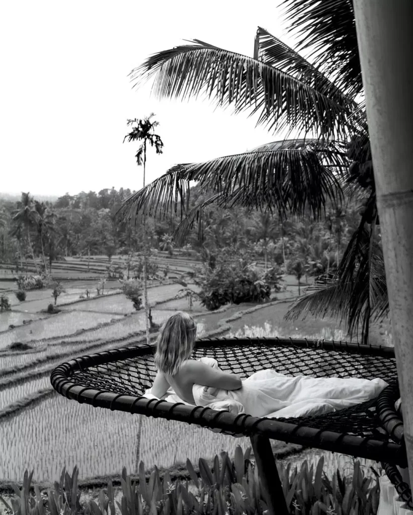 A solitary figure in a white dress relaxes on a net over an extensive landscape of **rice paddies** in this monochrome photograph. The image evokes the sense of **peace and quiet reflection** found when slowing down. Alt: To fall in love with Bali again, consciously uncouple from the algorithm. This quiet moment in the rice fields shows how the answer to is Bali overrated lies in seeking imperfect, unforgettable moments, not perfect photos.