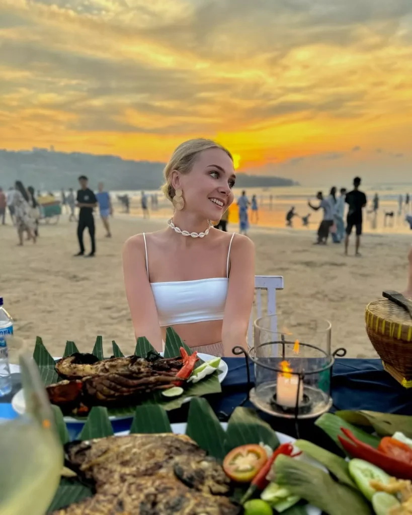 A tourist enjoying a Jimbaran sunset dinner on the beach. A smiling blonde woman wearing a white top and a seashell necklace is seated at a table with a feast of grilled seafood and a candle lantern, against the backdrop of a vibrant orange sunset and a crowded beach. This is a common destination for Bali Airport to Jimbaran Transfer.