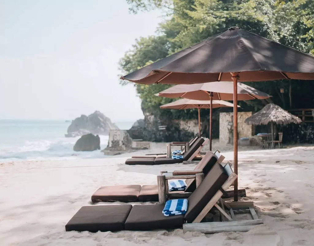 A serene beach scene in South Bali, featuring a row of wooden sun loungers with brown cushions and large brown umbrellas (parasols) on soft, light-colored sand. In the background, ocean waves break near large rock formations and green foliage on the cliffside. A striped blue and white towel is visible on one of the empty loungers, inviting relaxation.