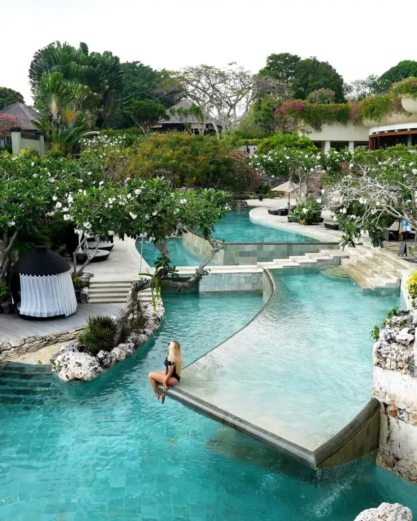 A stunning photo of the multi-tiered infinity pools and tropical gardens at Ayana Resort Bali in Jimbaran. A blonde woman in a black bikini is seated at the edge of a unique, curved pool section, overlooking the turquoise water and lush surroundings. This image represents the "Jimbaran Premium" luxury destination, which influences the high perceived cost of an airport transfer to Ayana due to the resort's exclusive, hilly location and luxury association.