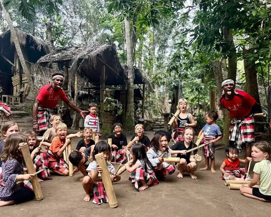 A group of diverse children joyfully participating in a traditional Balinese music or play activity outdoors, led by two local men in sarongs. Their genuine smiles and engagement directly challenge the notion that Bali is boring, showcasing the island's vibrant, authentic connections.