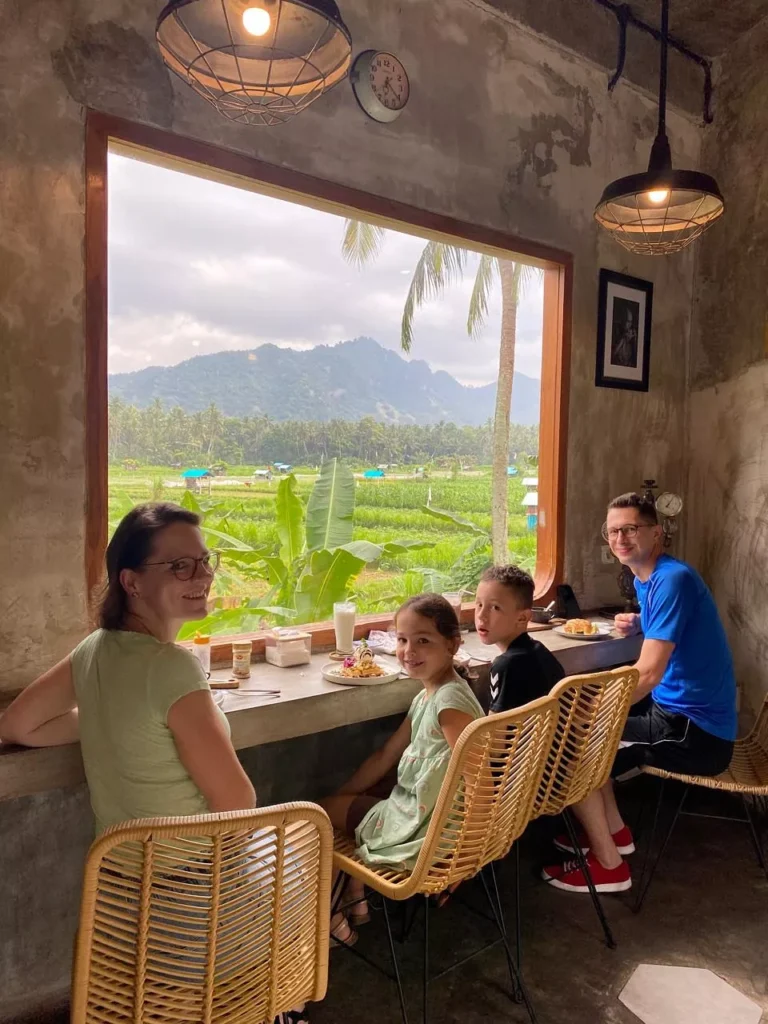 A family of four (two adults and two children) sitting at a counter inside a cafe with a large window overlooking a lush green landscape of rice fields and tropical mountains. The family is enjoying a meal. The alt text incorporates the user query: Is Ubud Worth Visiting?