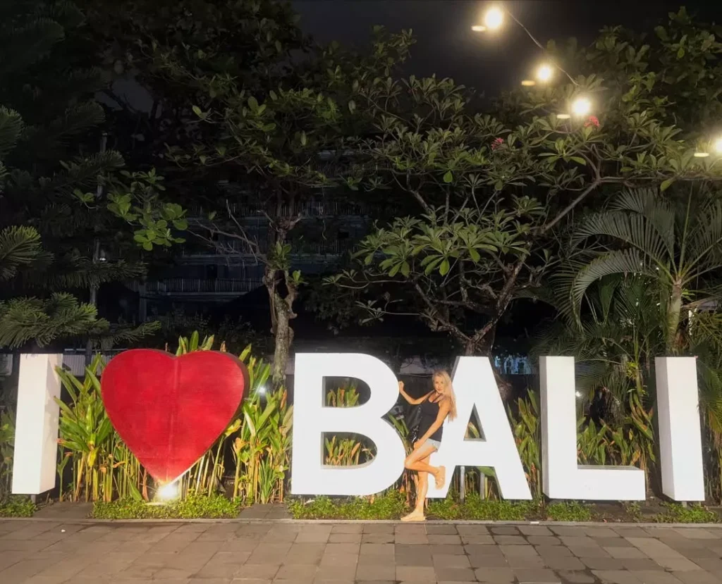 Woman posing in front of the large 'I ❤️ BALI' sign at night after arriving via the port, highlighting the need for a reliable Benoa Cruise Port Transfer.