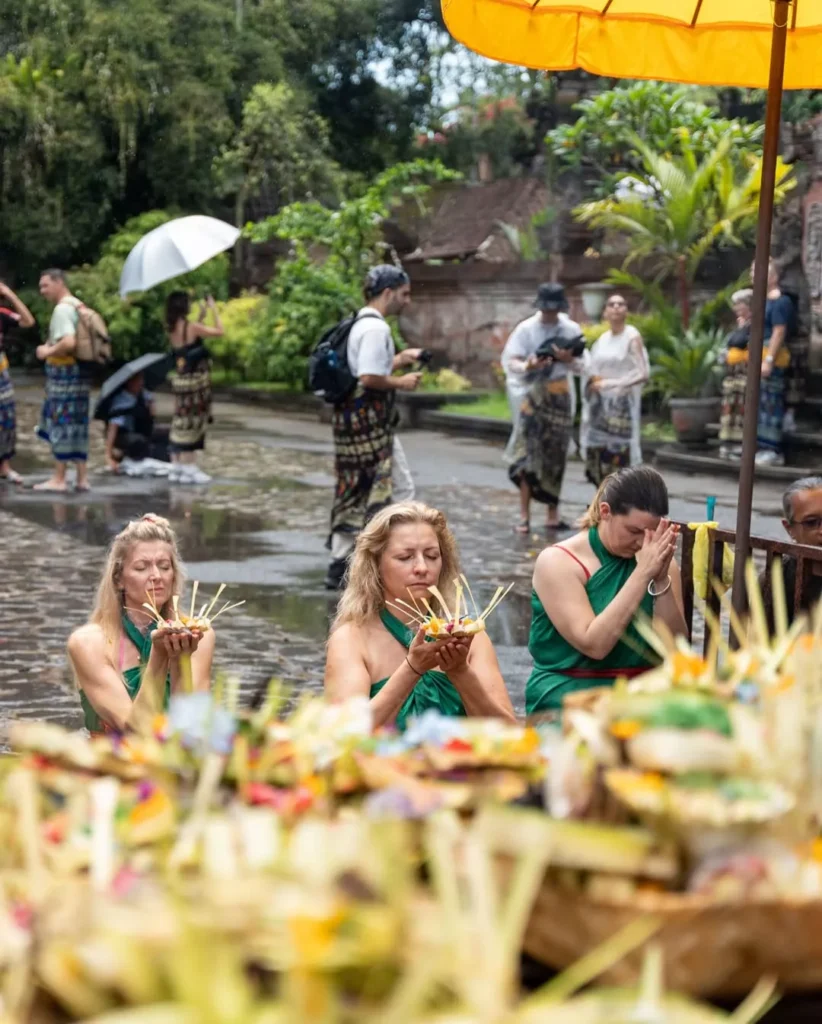 Tourists or participants, including three blonde women in green sarongs, performing a Hindu Balinese blessing or purification ritual (Melukat) in a temple courtyard, kneeling in shallow water and holding offerings. The foreground is blurred with traditional Balinese offerings (Canang Sari).