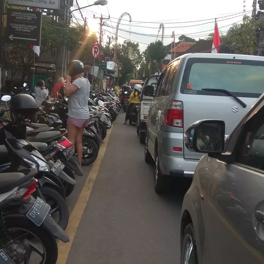 Ubud Traffic Jam on a busy street. The street is heavily congested with a long line of parked motorbikes (scooters) on the left, a silver van and other cars blocking the road, and overhead electrical wires visible. A tourist wearing a helmet and shorts is standing next to the parked bikes on the left. This highlights common Ubud Street Congestion issues.
