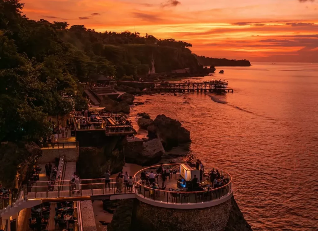 A dramatic sunset view over a rugged coastline, featuring a cliffside establishment with multiple tiered wooden decks and circular dining areas built onto the rocks. The setting sun casts a vibrant orange and red glow across the sky and the ocean water below. Guests are visible enjoying drinks and views at the popular oceanfront venue.
