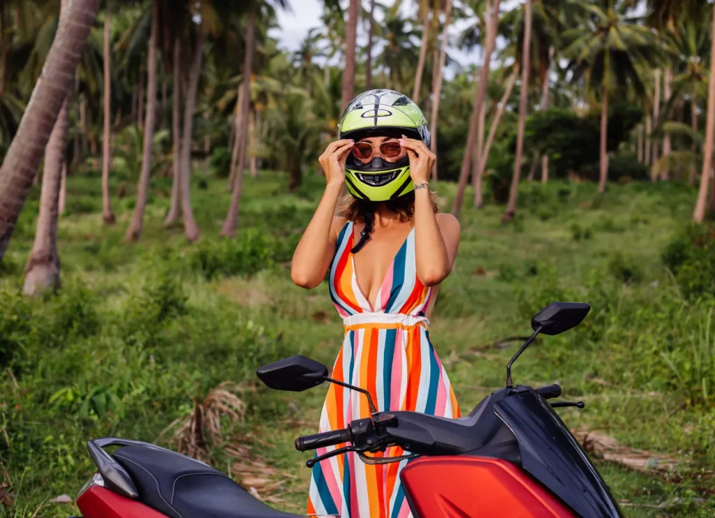A foreign woman adjusts her lime-green helmet while standing next to a red scooter in a palm forest, illustrating an essential method of transport in Bali.