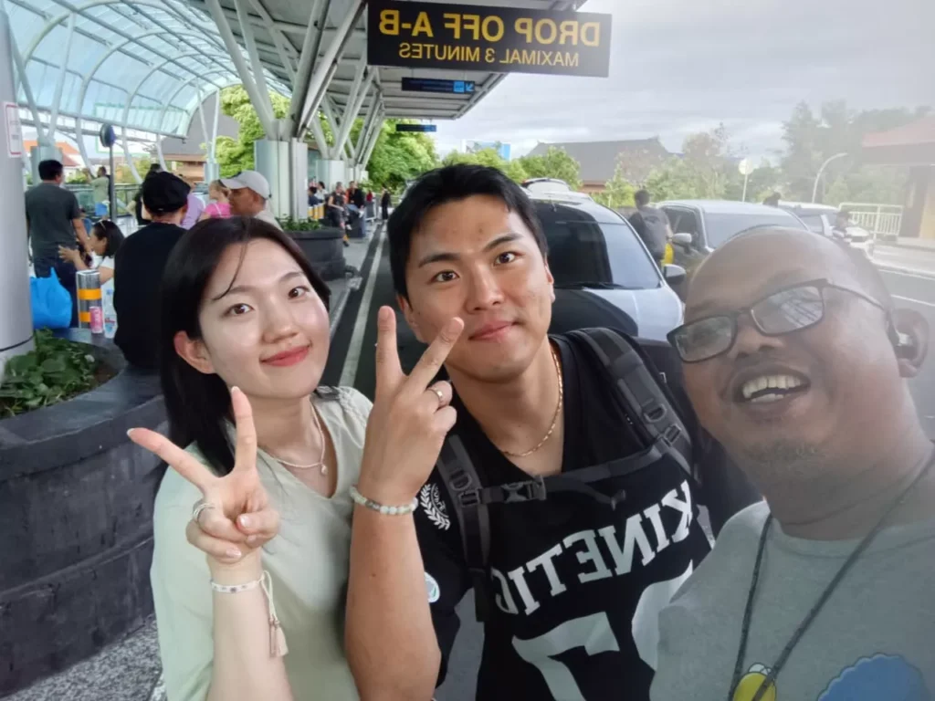 Hey Bali Team! - A selfie of a smiling couple and a local man (possibly a taxi driver or transfer service) at the Drop Off A-B area of an airport terminal. The sign above the group is mirrored and reads "DROP OFF A-B - MAX 3 MINUTES". The couple is holding up peace signs.