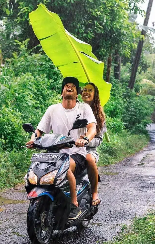 A pair of foreign tourists riding a motorbike in Bali during the rain while holding banana leaves to act as an umbrella.